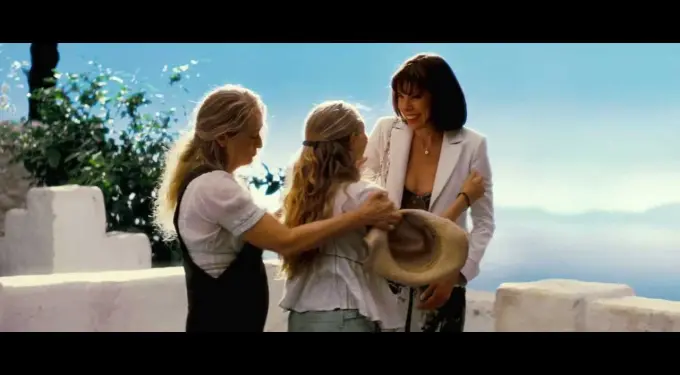 Three women, one holding hat, warmly greeting outside near white stone