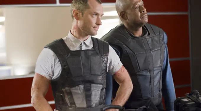 two men in tactical vests stand guarding equipment indoors