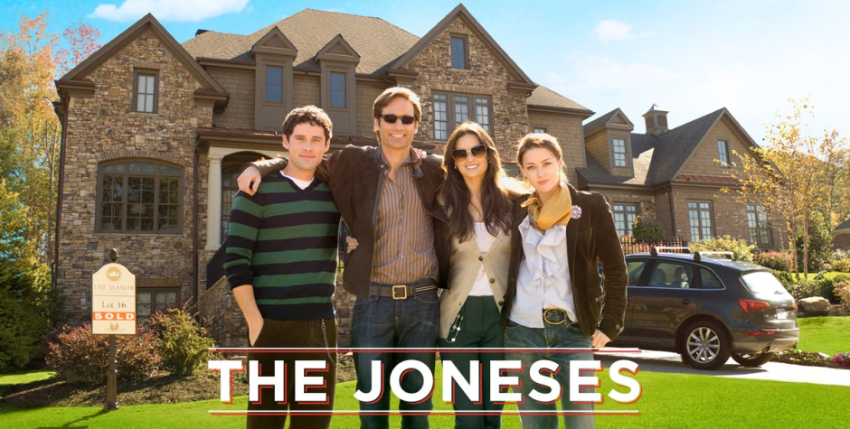 Four people stand smiling outside large house with The Joneses title