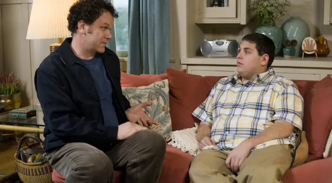 Two men sitting on couch, one gesturing, talking in living room