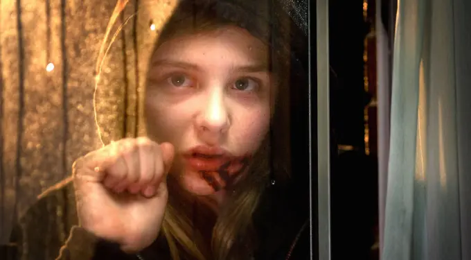 Woman with blood on mouth looks out window in dark scene