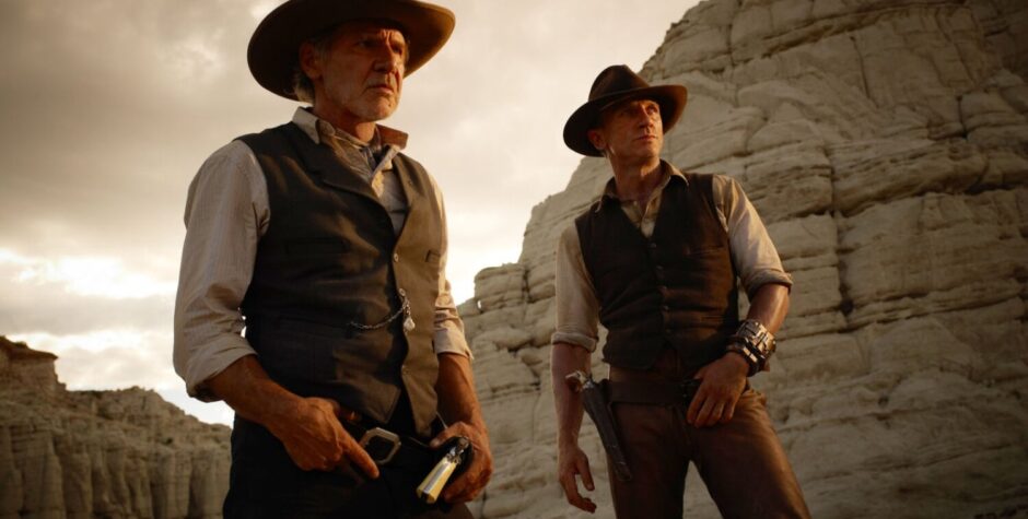 Two men in cowboy hats and vests stand in desert with guns