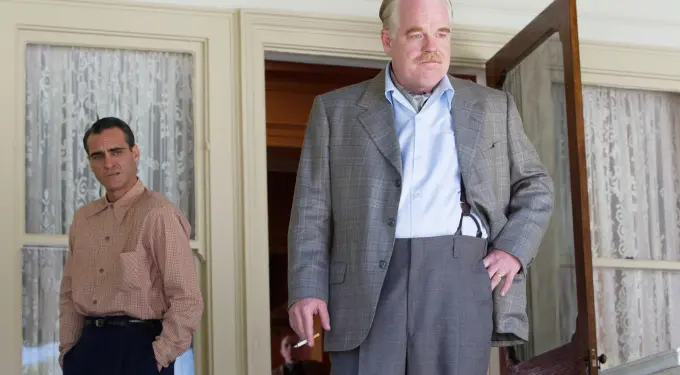 Two men in vintage outfits stand by a door and lace curtains indoors
