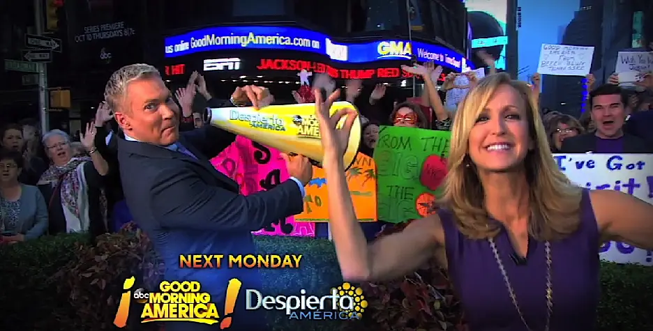 Hosts promote Good Morning America and Univision’s Despierta America to crowd with signs.
two morning show hosts pose with crowd and signs behind them