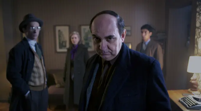 Man in dark coat in foreground with three people behind him in vintage room