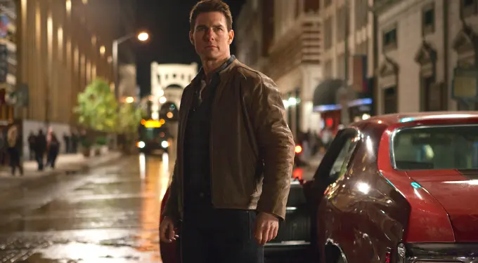 Man in brown jacket stands beside red car on wet street at night