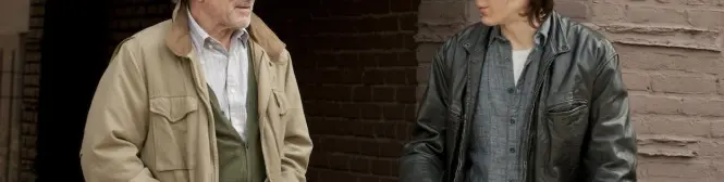 Two men talk outside near brick walls, one in leather jacket, one in tan coat