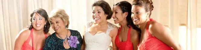 Five women smile together, one wearing a wedding dress indoors