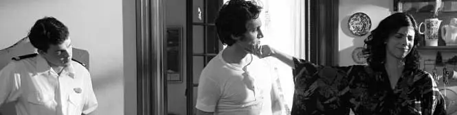 two men and a woman talk inside a kitchen in black and white