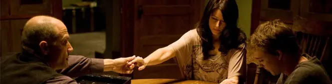 Three people hold hands across a dimly lit table in tense scene