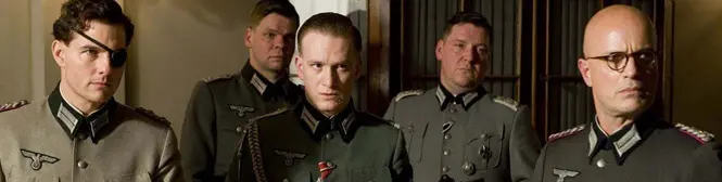 Four men dressed as Nazi officers stand indoors with serious faces