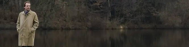 Man in tan coat standing by quiet lake with trees behind