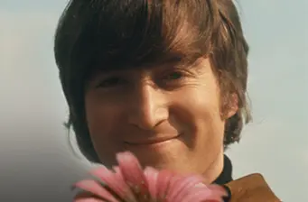 Young John Lennon smiles holding a pink flower outdoors