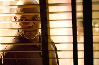 man looking through slatted window blinds with serious expression