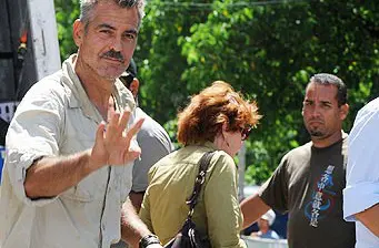 man waves at camera outdoors with people walking behind