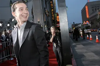 Shia LaBeouf smiling in a suit on street with photographers behind