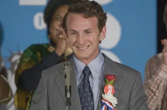 Man in suit with rose boutonniere speaks at microphone, smiling