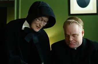 two hooded figures, one woman with glasses, one man dressed as priest