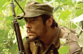 man in military outfit crouches holding a rifle alert in bushes