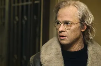 man with glasses and fur-collared coat looks to right indoors