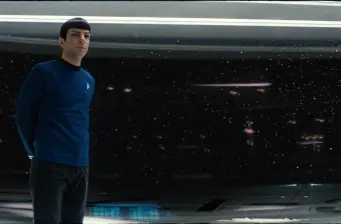 Star Trek character in blue uniform stands on starship bridge