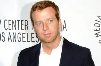 Man in suit with light hair stands in front of white media wall