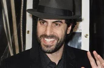 Sacha Baron Cohen smiling in black hat and suit