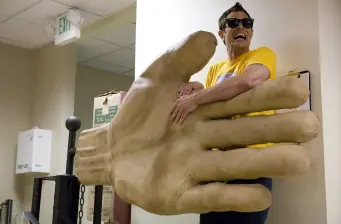 Man in sunglasses holds oversized foam hand indoors with surprised expression