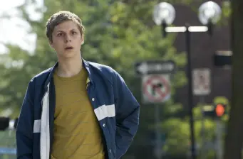 Michael Cera stands outside in blue jacket and yellow shirt