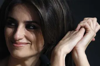 Penélope Cruz smiling with hands near face against a dark background