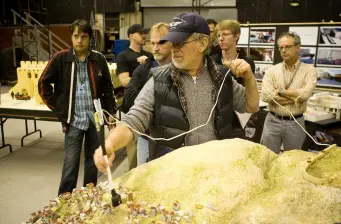 Steven Spielberg directs while holding model, men watch behind him