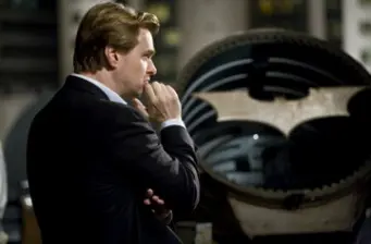 Man in suit stands pensively near Batman symbol in dark setting