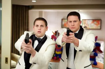 Two men in white tuxedos aim guns in indoor room