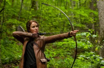 Woman aiming bow and arrow in forest wearing brown jacket