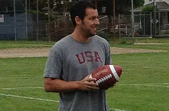Adam Sandler in gray USA shirt holding football on field