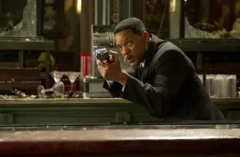 Man in black suit holds gun behind counter in tense scene