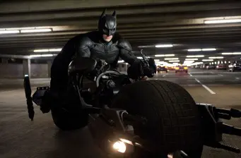Batman rides the Batpod motorcycle in a dimly lit parking garage