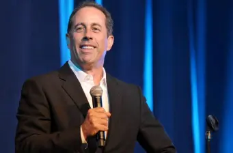 Jerry Seinfeld holds microphone, smiling during live event
