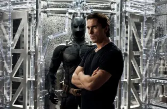 Christian Bale as Bruce Wayne stands before Batman suit display case