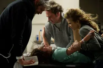 Two adults restrain a young girl lying on the floor in movie scene