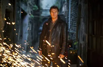 Liam Neeson in leather jacket standing with sparks flying around