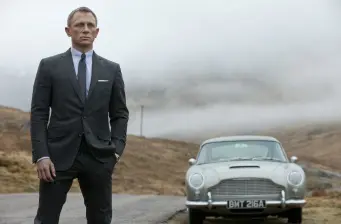 Daniel Craig in suit stands by vintage car in foggy landscape