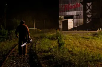 Man with chainsaw walks on railroad tracks at night near warehouse