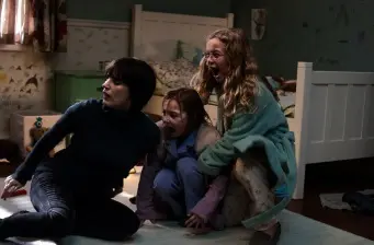 three frightened girls crouch near a bed in a dim bedroom scene