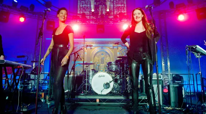 Two women singing on stage with bright purple and red lights