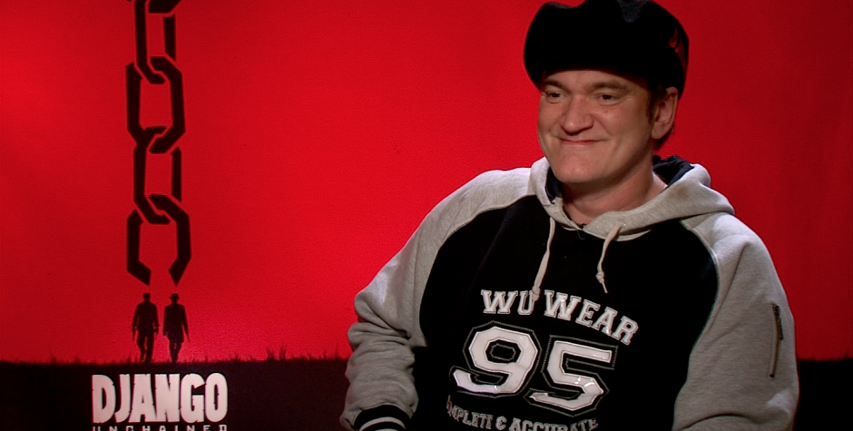 Quentin Tarantino smiling in casual wear with Django Unchained poster behind