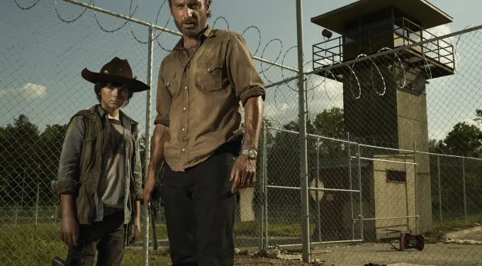 Two men stand outside barbed wire prison fence with watchtower