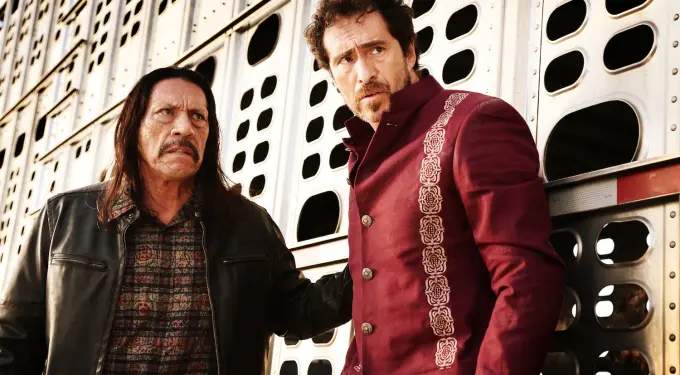 Two tense men stand by metal truck wall, one in embroidered jacket