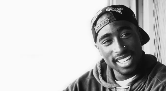 Close-up black and white photo of Tupac Shakur smiling in a backwards cap