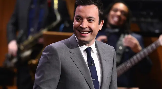 Jimmy Fallon smiling on stage wearing grey suit and navy tie